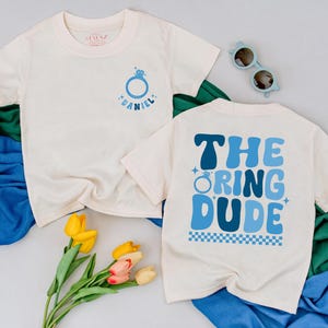 May include: Two off-white t-shirts. One has a blue ring graphic with the name "Daniel". The other shirt has the text "THE RING DUDE" in blue. The shirts are on a light gray surface with blue and green fabric.