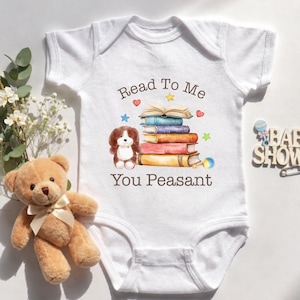 Puede incluir: Body de bebé blanco con el texto "Read To Me You Peasant" y un dibujo de libros, un cachorro y estrellas. Un osito de peluche y flores están al lado del body. Un letrero de madera que dice "Baby Shower" también está en la imagen.
