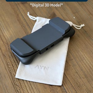 Puede incluir: Un accesorio de modelo 3D digital Odin 2 Portal Cover en color negro con acabado mate, que presenta agarres laterales contorneados y un panel central rectangular. Se muestra junto a una bolsa de tela blanca con cordón y el logo AYN en relieve.