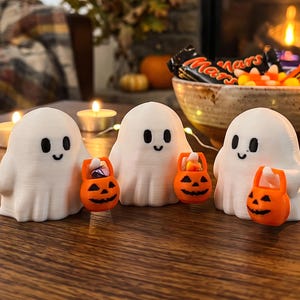 May include: Three white ghost figurines with smooth, rounded shapes and simple black eyes and mouths. Each ghost is holding a small, bright orange plastic jack-o'-lantern bucket filled with tiny pieces of colorful candy for Halloween.