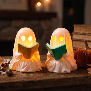May include: Two white ghost-shaped figurines with glowing yellow eyes, each holding an open book. One ghost holds a yellow book, and the other holds a green book. The figures are illuminated from within, casting a warm light on their surroundings.