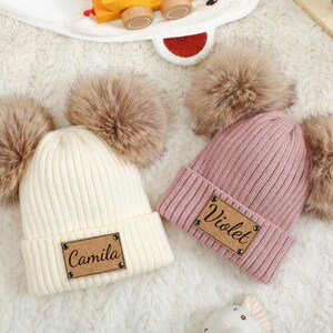 Può includere: Due berretti lavorati a maglia con pompon in finta pelliccia. Uno è color crema con il nome "Camila" su una toppa marrone. L'altro è rosa con "Violet" su una toppa simile. Entrambi sono su una superficie bianca e soffice.
