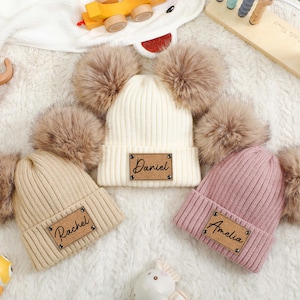 May include: Three ribbed knit beanies with faux fur pom-poms. One is cream, one is tan, and one is pink. Each beanie has a rectangular patch with a name: "Daniel", "Rachel", and "Amelia".
