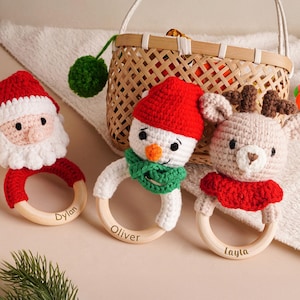 Op de afbeelding: Gehaakte kerst baby rammelaars. Ze tonen een kerstman, een sneeuwpop en een rendier, elk met een houten ring als basis. De kerstman heeft een rode muts en baard, de sneeuwpop een groene sjaal en het rendier een rode kraag. De rammelaars zijn gepersonaliseerd met namen.