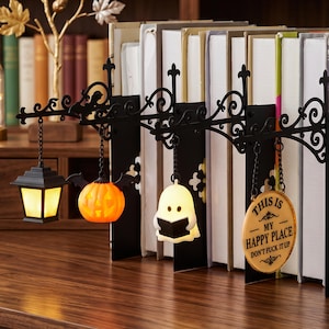 May include: A black metal bookend set featuring an ornate scrollwork top bar with four hanging charms: a lantern, a jack-o'-lantern, a ghost reading a book, and a round sign that reads "THIS IS MY HAPPY PLACE DON'T FUCK IT UP".