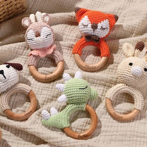 May include: A collection of handmade crochet baby rattles in various animal shapes, including a dog, fox, deer, giraffe, and dragon. Each rattle features a wooden ring and is crafted in soft, neutral colors with embroidered details. The rattles are designed for infants.