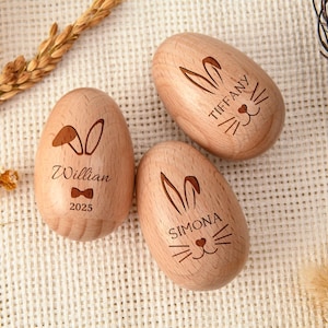 Puede incluir: Tres huevos de Pascua de madera con caras de conejito grabadas y los nombres: William, Tiffany y Simona. El huevo de William incluye una pajarita y el año 2025. Los huevos son de color marrón claro y están sobre una superficie blanca texturizada.