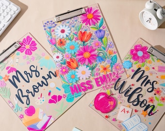 May include: Three clear acrylic clipboards featuring colorful floral illustrations and school-themed graphics. Two clipboards are personalized with the names Mrs. Brown and Mrs. Wilson in black script, while one features the name MISS EMILY in pink.