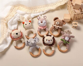 May include: A collection of handmade crochet baby rattles in various animal designs, including a lion, elephant, bear, deer, and rabbit. Each rattle features a wooden ring and is crafted in soft, neutral colors. The rattles are arranged on a light-colored surface.