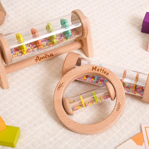 May include: Two wooden baby rattles with clear cylindrical chambers filled with colorful small beads. One rattle is a horizontal cylinder on a stand engraved with the name Amira, and the other is a circular ring rattle engraved with the name Matteo.