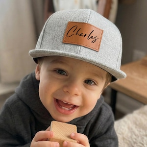 May include: A gray flat-brimmed hat with a brown leather patch that reads "Charles" in a cursive font. The hat is worn by a smiling child wearing a gray hooded sweatshirt.