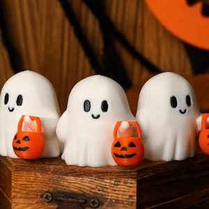 May include: Three small white ghost figurines with rounded shapes and smooth surfaces. Each ghost has simple black oval eyes and a small smile, and holds a miniature bright orange plastic pumpkin bucket with a carved jack-o'-lantern face.