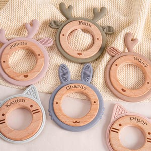 Puede incluir: Una colección de juguetes para la dentición del bebé en varios colores pastel, incluyendo rosa, verde y azul. Cada juguete presenta un anillo de madera en el centro con una forma de animal de silicona, como un ciervo, un gato o un conejo. Los nombres están grabados en cada juguete.