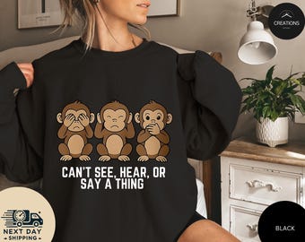 May include: Black sweatshirt featuring three cartoon monkeys in a row, each with a different gesture: covering eyes, ears, and mouth. The text "CAN'T SEE, HEAR, OR SAY A THING" is below the monkeys. The sweatshirt is a crew neck style.