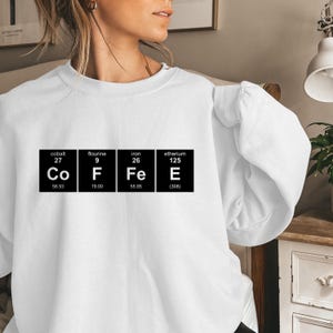 May include: White sweatshirt with a graphic of the periodic table elements spelling out "COFFEE".