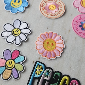 Könnte beinhalten: Gestickte Aufnäher mit positiven Botschaften und floralen Designs. Enthält Aufnäher mit den Worten "Love" und "Peace" sowie Smiley-Blumen-Designs in verschiedenen Farben. Andere Aufnäher sagen "Be Kind" und "Always Stay Positive".