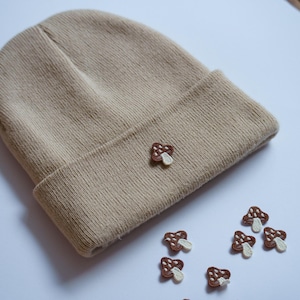 Op de afbeelding: Een beige beanie met een geborduurd bruin paddenstoel op de voorkant. Er zijn verschillende bruine paddenstoel patches rond de beanie verspreid.