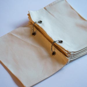 Op de afbeelding: Een stapel blanco witte stofpagina's gebonden met zilveren ringen. De pagina's zijn dubbelgevouwen en de omslag is open.