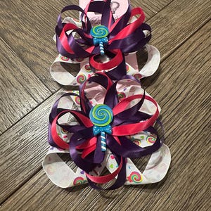 Pode incluir: Dois laços decorativos para cabelo. Cada laço apresenta camadas de fitas roxas, rosas e brancas, com um enfeite em forma de pirulito no centro. Os laços estão sobre uma superfície de madeira.