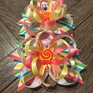 Pode incluir: Dois laços de cabelo em camadas com tema de doces. Os laços são feitos de fitas de cetim amarelas, rosas e brancas. Cada laço tem um enfeite de pirulito vermelho e amarelo no centro. Os laços estão em um fundo listrado.