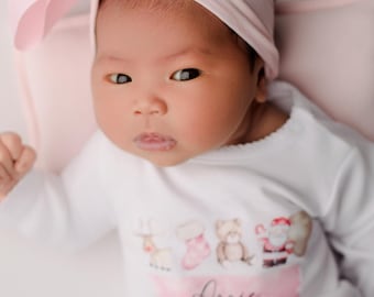 May include: A newborn baby wearing a white onesie with a pink bow headband and a matching pink pillow. The onesie has a pink banner with the name "Arrie" and Christmas-themed graphics.