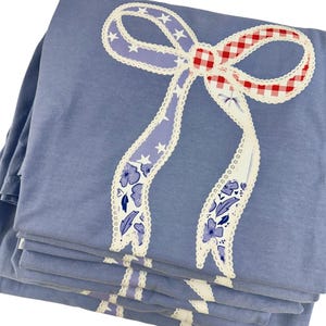 May include: A stack of blue sheets with a large bow design. The bow features a red and white gingham center, lavender ribbons with white stars, and white lace trim. Floral and leaf patterns decorate the ribbon.