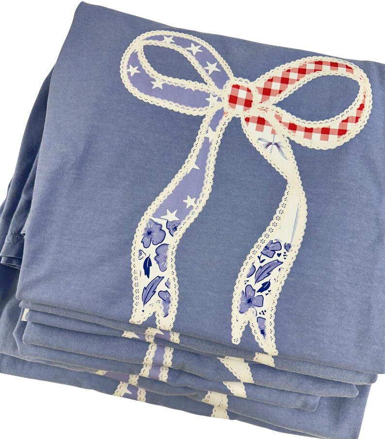 May include: A stack of blue sheets with a large bow design. The bow features a red and white gingham center, lavender ribbons with white stars, and white lace trim. Floral and leaf patterns decorate the ribbon.