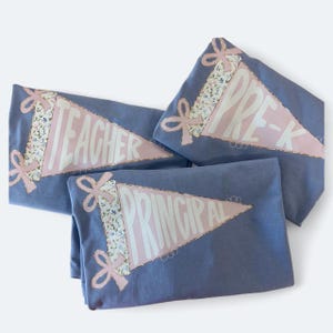 May include: Three blue pennant-shaped shirts with the words "TEACHER", "PRE-K", and "PRINCIPAL" in white. Each shirt has a pink pennant with a floral pattern and pink bows.