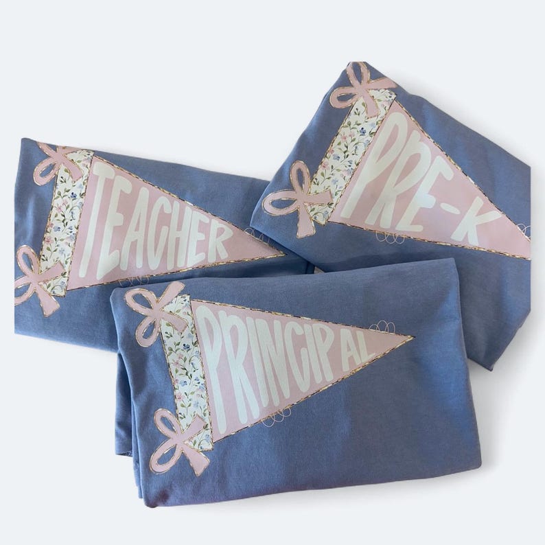 May include: Three blue pennant-shaped shirts with the words "TEACHER", "PRE-K", and "PRINCIPAL" in white. Each shirt has a pink pennant with a floral pattern and pink bows.