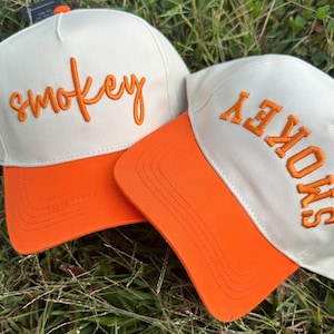 Può includere: Due cappellini da baseball con corona color crema e visiera arancione. La parola "smokey" è ricamata in arancione sulla parte anteriore di ogni cappellino. I cappellini sono appoggiati sull'erba verde.