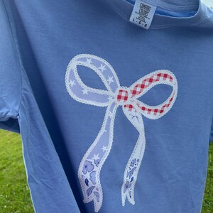 May include: A light blue t-shirt featuring a large bow design. The bow has a red and white gingham center, white lace trim, and light blue ribbons with star and floral patterns. The shirt has a Comfort Colors tag.