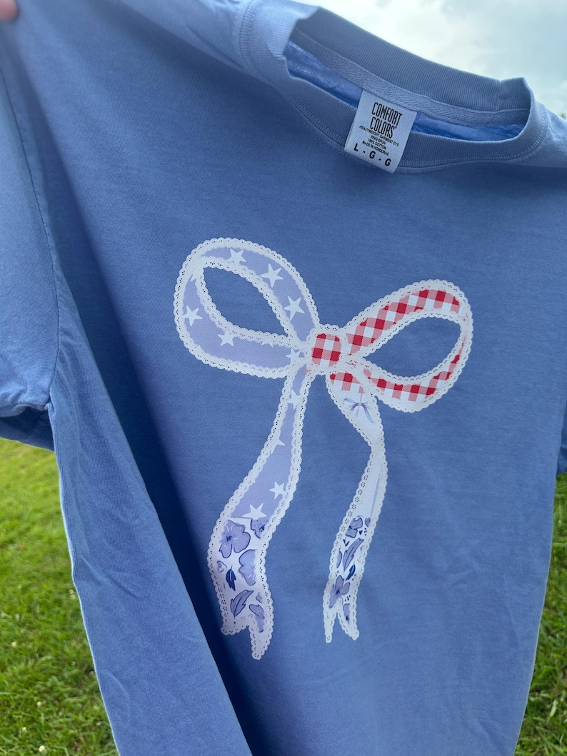 May include: A light blue t-shirt featuring a large bow design. The bow has a red and white gingham center, white lace trim, and light blue ribbons with star and floral patterns. The shirt has a Comfort Colors tag.