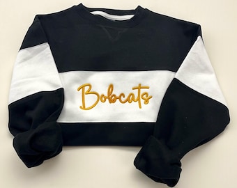 Puede incluir: Sudadera negra, blanca y dorada con la palabra "Bobcats" bordada en el pecho. La sudadera tiene un diseño de bloques de color con mangas negras y una franja blanca en el pecho.