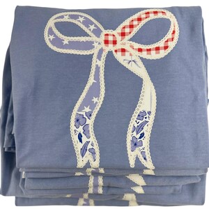 May include: A stack of folded blue fabric items, possibly shirts or sheets, featuring a large bow design. The bow has red and white gingham, purple ribbons with white stars, and floral patterns, all outlined with white lace.