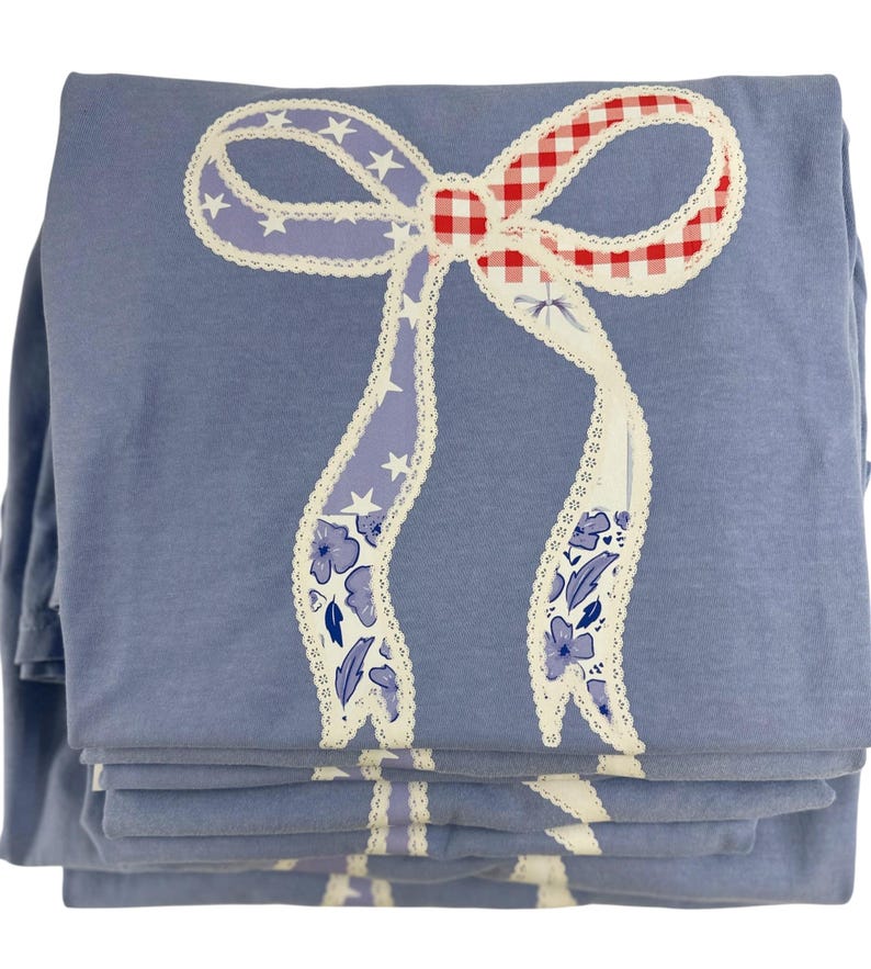 May include: A stack of folded blue fabric items, possibly shirts or sheets, featuring a large bow design. The bow has red and white gingham, purple ribbons with white stars, and floral patterns, all outlined with white lace.