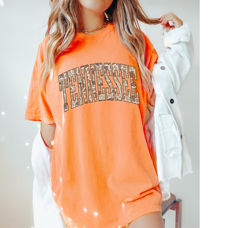 Puede incluir: Camiseta naranja con la palabra "TENNESSEE" en un estampado de camuflaje en el pecho. La camiseta tiene mangas cortas y cuello redondo. La camiseta es de color naranja liso.