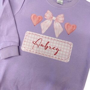 Peut inclure: Un sweat-shirt lavande avec un nœud rose, deux cœurs roses et un rectangle en vichy blanc et rose avec le nom "Aubrey" écrit en cursive.