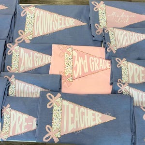 May include: A collection of light blue and pink t-shirts. Each shirt features a pennant design with text such as "TEACHER", "SCHOOL PSYCH", "2ND GRADE", and "3RD GRADE". The pennants have a floral ribbon design.