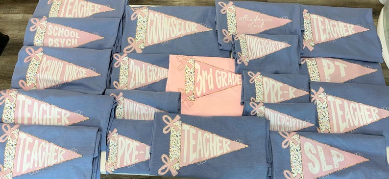 May include: A collection of light blue and pink t-shirts. Each shirt features a pennant design with text such as "TEACHER", "SCHOOL PSYCH", "2ND GRADE", and "3RD GRADE". The pennants have a floral ribbon design.