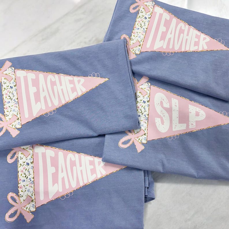 May include: Several blue t-shirts with a pennant design. The pennants are pink with gold trim and feature the word "TEACHER" or "SLP" in white. The pennants have a floral pattern and are decorated with pink bows.