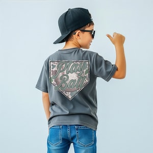 Può includere: Una maglietta grigia con una grafica a tema baseball sul retro. La grafica presenta le parole "Play Ball" in un font stilizzato, all'interno di una forma di base di baseball. La maglietta è abbinata a jeans blu e un berretto da baseball nero.