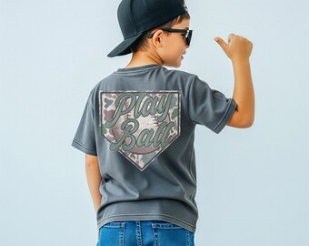 Puede incluir: Una camiseta gris con un gráfico de béisbol en la espalda. El gráfico presenta las palabras "Play Ball" en una fuente estilizada, dentro de una forma de plato de home de béisbol. La camiseta se combina con vaqueros azules y una gorra de béisbol negra.