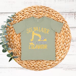 Può includere: T-shirt verde salvia con grafica di escavatore giallo e la scritta "See Ya Later Excavator". La maglietta è esposta su un sottobicchiere circolare intrecciato con fogliame verde.