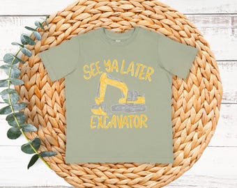 Puede incluir: Camiseta verde salvia con un gráfico de excavadora amarilla y el texto "See Ya Later Excavator". La camiseta se muestra sobre un salvamanteles circular tejido con follaje verde.