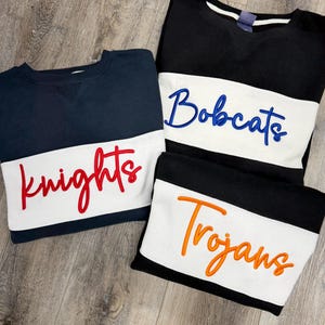 May include: Three crewneck sweatshirts in navy, black, and black. Each has a white horizontal stripe across the chest with embroidered team names: "Knights" in red, "Bobcats" in blue, and "Trojans" in orange.