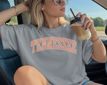 May include: A gray t-shirt with the word "TENNESSEE" in orange block letters. The shirt has a crew neck and short sleeves. The person in the image is wearing sunglasses and holding a beverage in a clear cup with a black lid and straw.