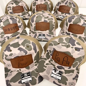 Può includere: Una collezione di otto cappellini da camionista mimetici con toppe in pelle marrone. Le toppe hanno diversi messaggi scritti a mano, tra cui "Daddy", "Dad" e "Pa Pa".