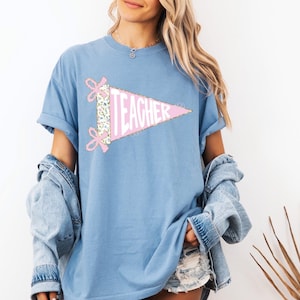 May include: A light blue t-shirt featuring a pink pennant with the word "TEACHER" in white. The pennant is decorated with floral fabric and pink bows. The shirt is part of a clothing collection.