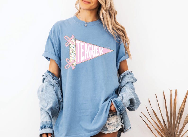 May include: A light blue t-shirt featuring a pink pennant with the word "TEACHER" in white. The pennant is decorated with floral fabric and pink bows. The shirt is part of a clothing collection.