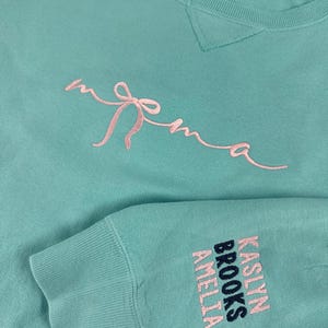 May include: A light teal sweatshirt with the word "mama" embroidered in pink script with a bow design. The sleeve features the names "Kaslyn Brooks Amelia" embroidered in pink and navy.